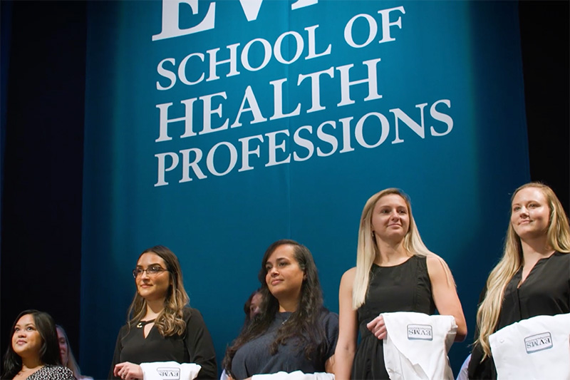 Group of students receive white coats on stage at ceremony standing in front of blue backdrop