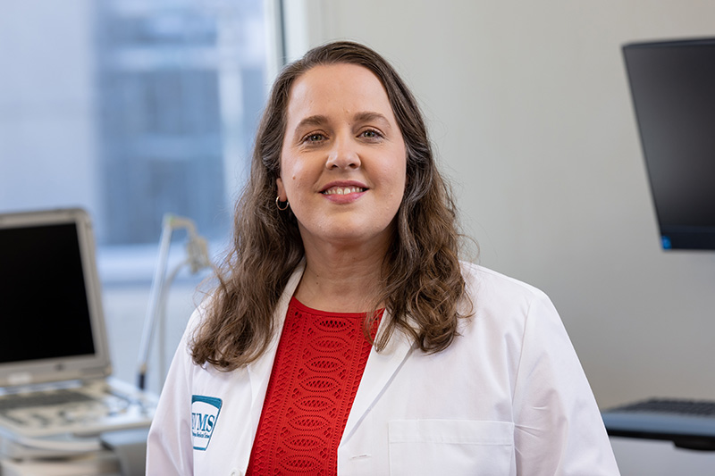 Dr. Morales has long wavy brown hair, is wearing a red sweater under a white medical coat in a clinical setting, and is smiling at the camera