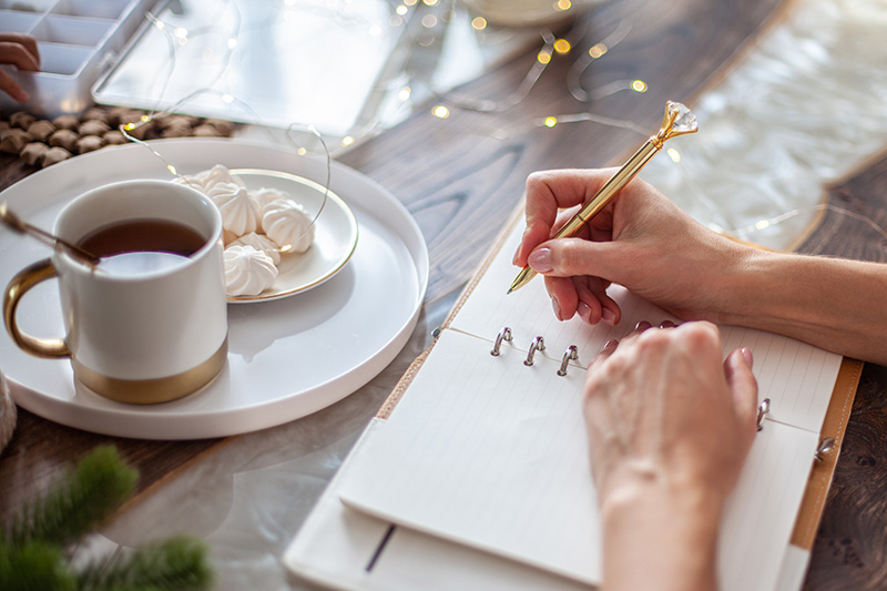 A woman writes in a blank notebook with tea and cakes on a table decorated with sparkly white lights, white and gold ribbon, and evergreen branch.