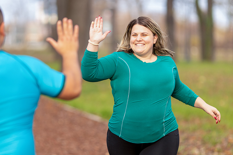 A happy overweight woman in teal blue sportswear top high-fives another runner while exercising