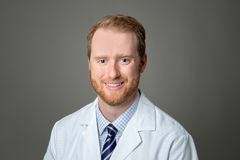 Dr. Whitlock has short strawberry blond hair, beard and mustache, is wearing a light blue dress shirt and tie under a white medical coat and is smiling at the camera
