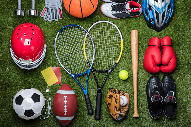 Various sports equipment lays on a green grass background. Includes football, soccer ball, baseball glove, softball, tennis racquets, bat, safety gear.