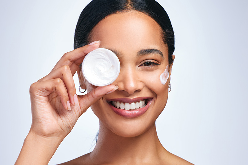 A smiling woman holds a small container of moisturizer as she applies it to her face.