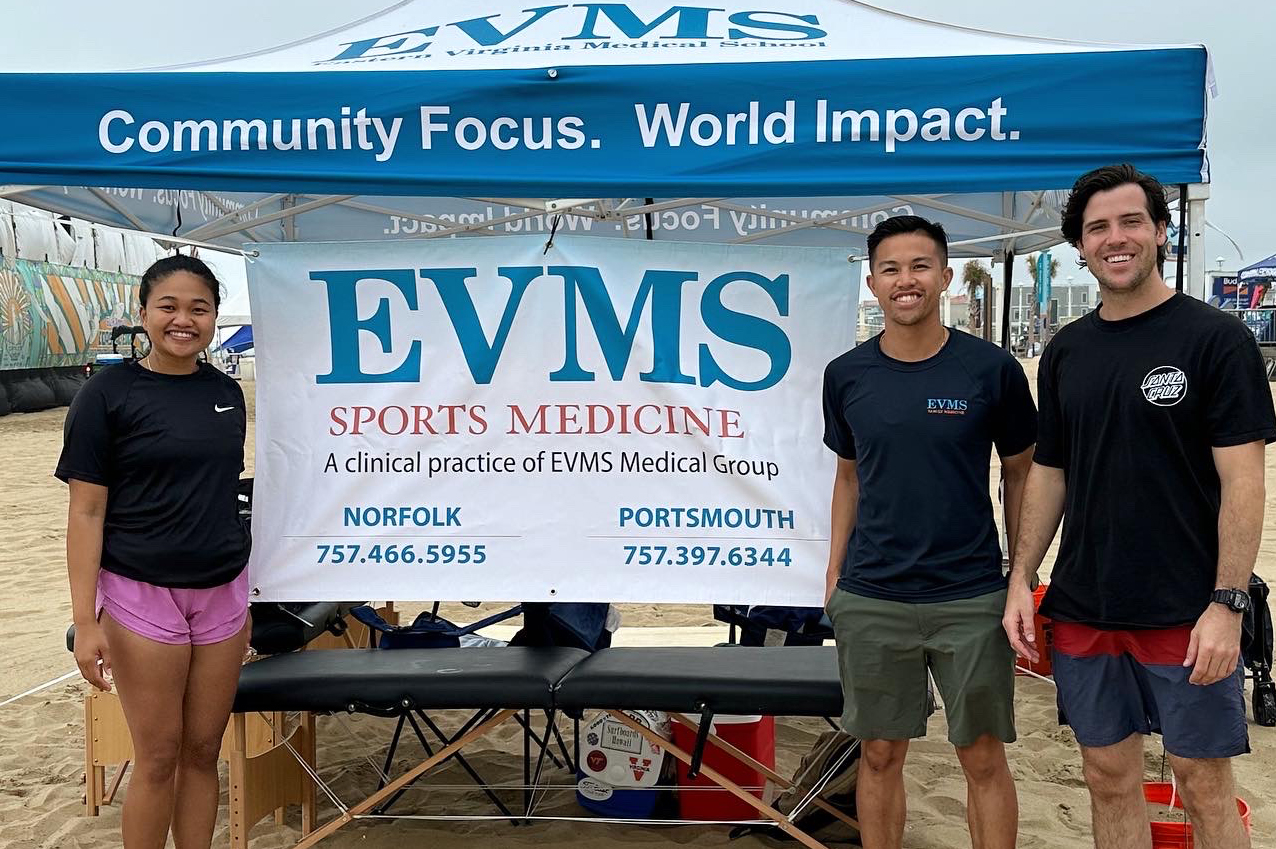 Two men and one woman wear shorts and t-shirts while standing on the beach in front of a sign that reads EVMS sports medicine community focus world impact