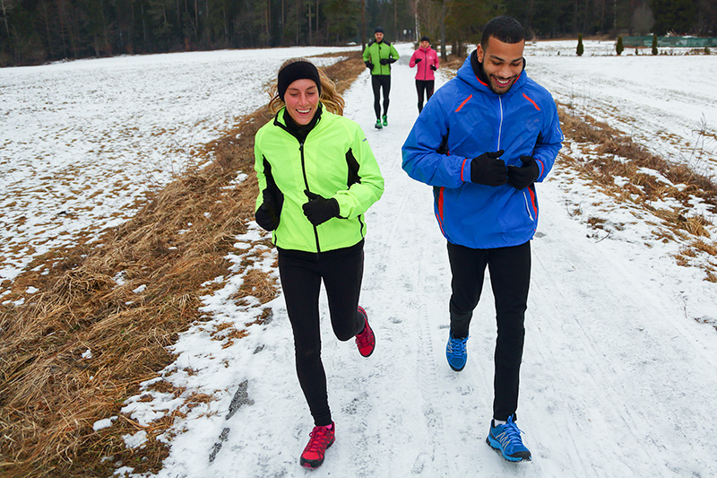 Four people dressed in bright winter clothing jog on a snow-covered dirt road. The two closest to the camera are smiling.